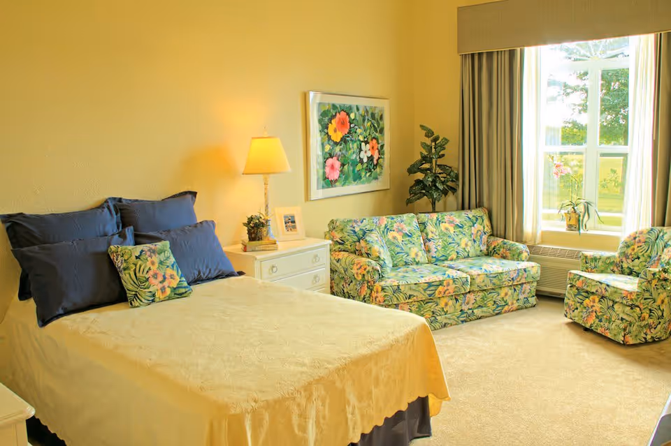 A cozy bedroom with a bed covered in a yellow bedspread and blue pillows. Next to the bed is a white nightstand with a lamp, a small plant, and a framed photo. Across from the bed is a floral-patterned sofa and matching armchair near a large window with beige curtains. A colorful floral painting hangs on the wall above the sofa, and a potted plant is placed in the corner.