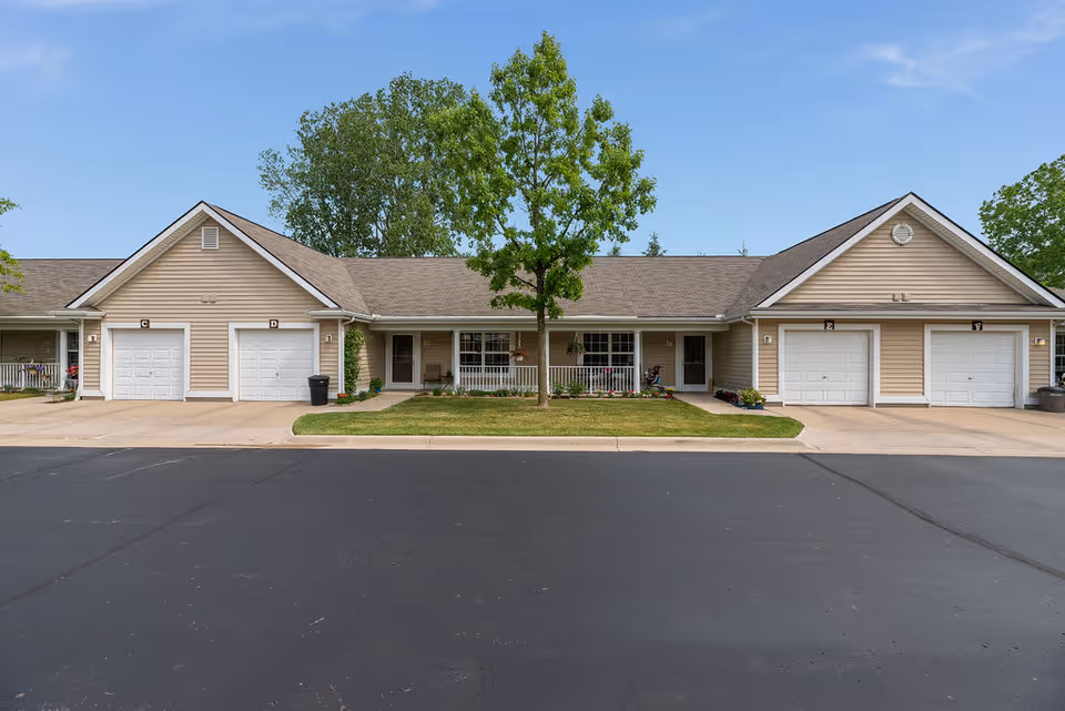 Exterior view of a single-story residential building with beige siding and four white garage doors labeled C, D, E, and F. There is a small lawn with a tree and a paved driveway in front of the building. The sky is clear with a few clouds.