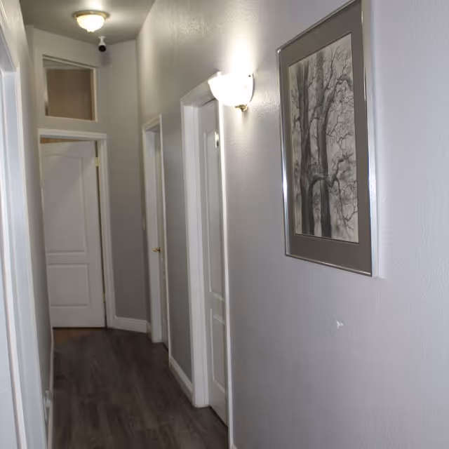 Narrow interior hallway with gray walls, wood flooring, multiple white doors, a framed tree print and a wall sconce.