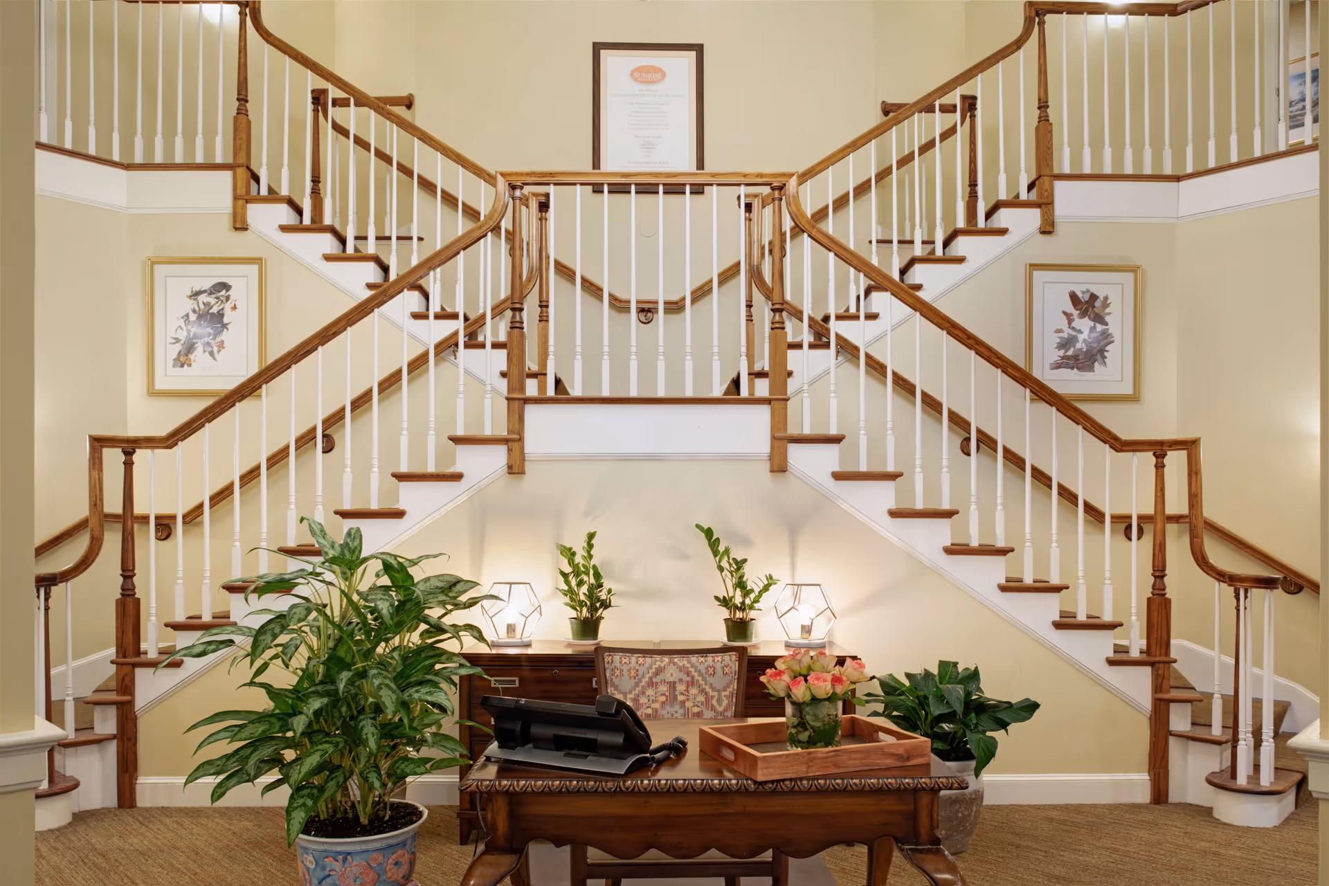Interior view of a senior living facility lobby area featuring a wooden double staircase with white balusters. Below the staircase is a wooden desk with a telephone, a wooden tray with a vase of pink roses, and a chair with patterned upholstery. There are several potted plants around the desk and two framed pictures on the walls beside the staircase. A framed mission statement is mounted on the wall above the staircase.