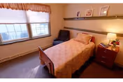 A small, cozy bedroom with a single bed covered in a peach-colored quilt. Next to the bed is a wooden nightstand with a lamp and a small plant. There is a dark armchair in the corner near a large window with blinds and a valance. Above the bed is a long shelf holding framed pictures and decorative items.