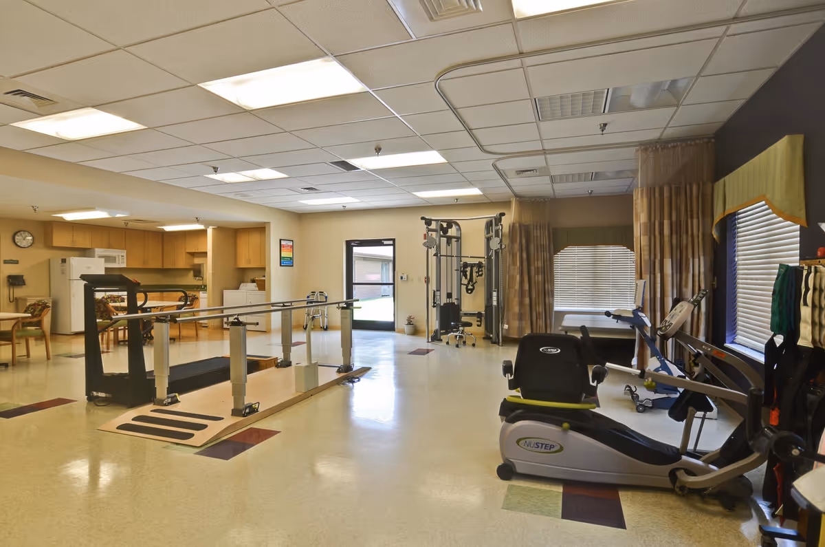 Well-lit therapy/exercise room with parallel walking bars, cardio machines, and a kitchenette area in the background.