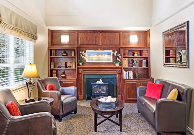 A cozy living room with two brown leather armchairs and a brown leather sofa, each adorned with colorful throw pillows. A wooden coffee table with decorative items sits in the center on a carpeted floor. Behind the seating area is a wooden built-in bookshelf and fireplace with a framed landscape painting above it. A table lamp is placed next to one of the armchairs near a window with blinds and a beige valance. A large mirror hangs on the wall above the sofa.