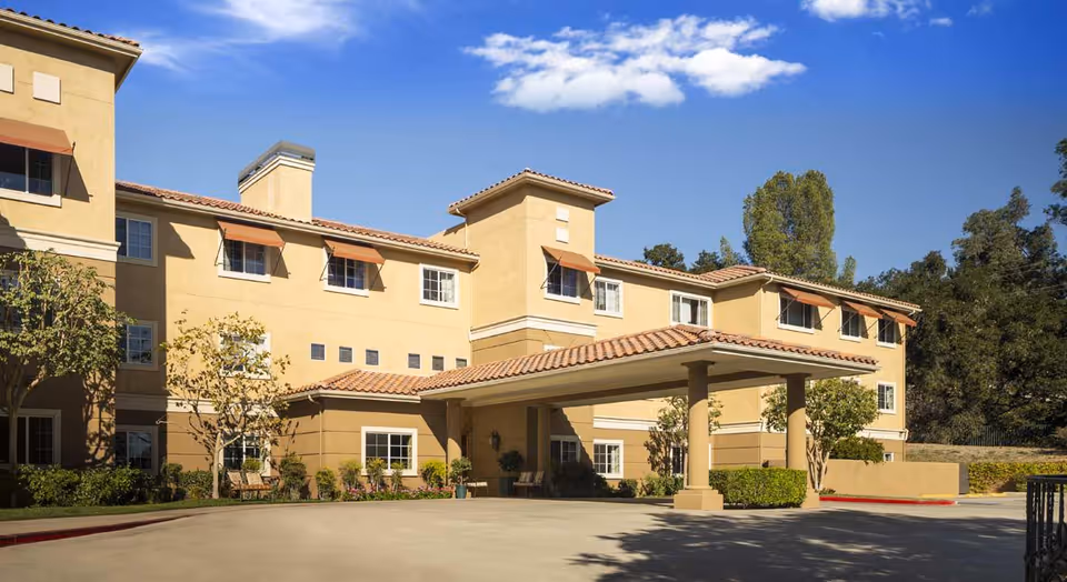 Exterior view of a three-story senior living facility building with beige walls, red-tiled roof, and multiple windows with awnings. The entrance has a covered driveway supported by columns, surrounded by trees and shrubs under a clear blue sky.