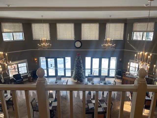 View from an upper level overlooking a spacious dining area with multiple tables and chairs arranged neatly. A decorated Christmas tree stands near large windows that let in natural light. The room features high ceilings with chandeliers and several windows with shades partially drawn. A clock is mounted on the wall above the windows.