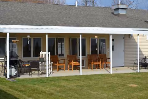 Covered porch with wooden chairs and a grill on a grassy exterior of a retirement facility.
