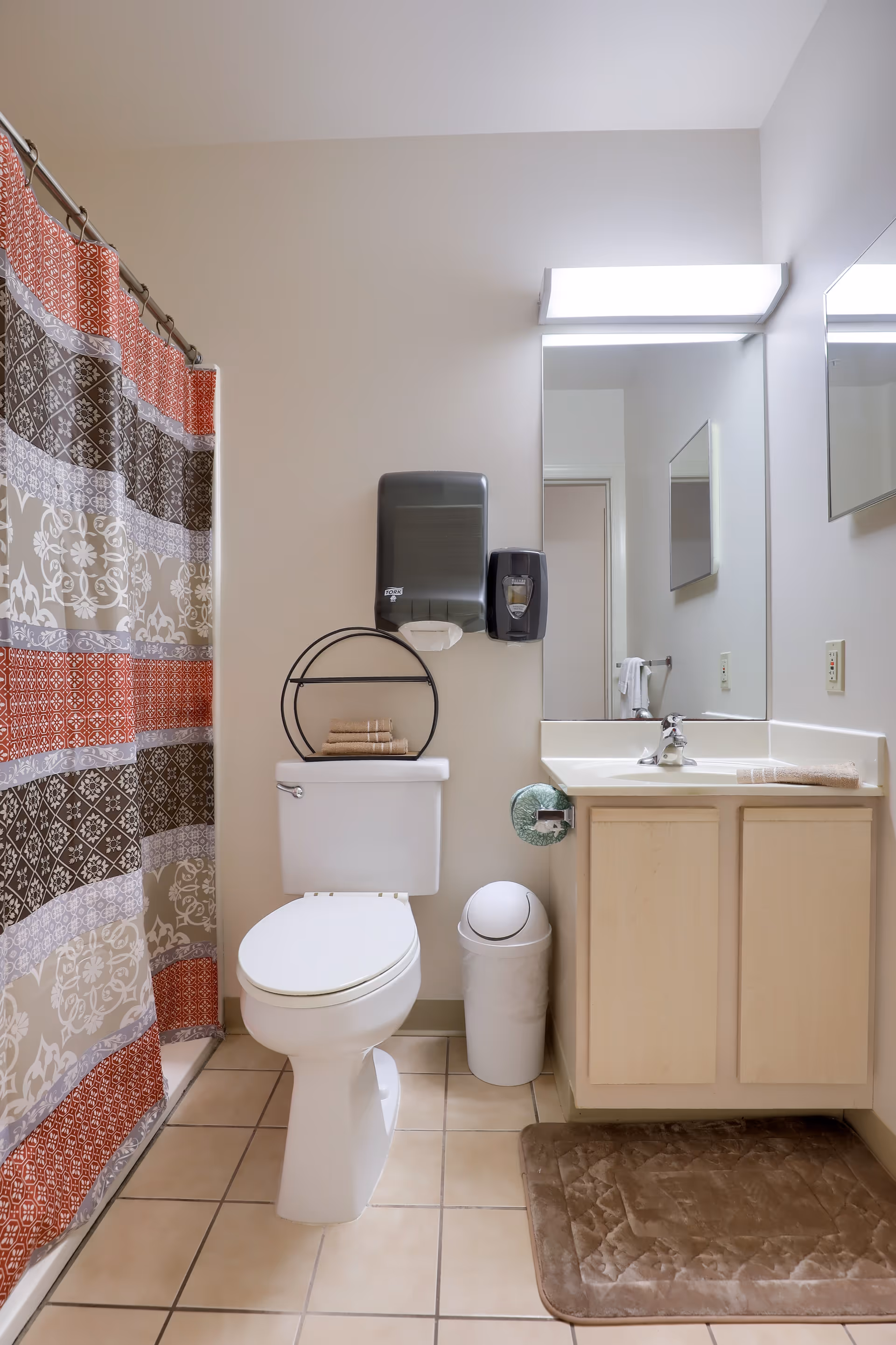 Compact bathroom featuring a toilet, sink vanity with mirror, wall-mounted dispensers, and a patterned shower curtain.