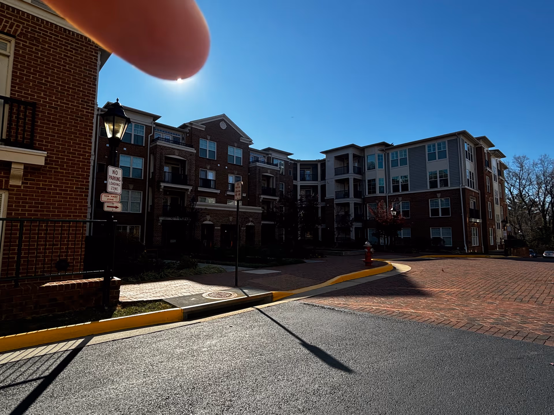 Exterior view of a multi-story brick and siding residential building under a clear blue sky. The building has multiple windows and balconies. There is a paved driveway and sidewalk with a streetlamp and a 'No Parking Loading Zone' sign. A finger partially covers the top left corner of the image.