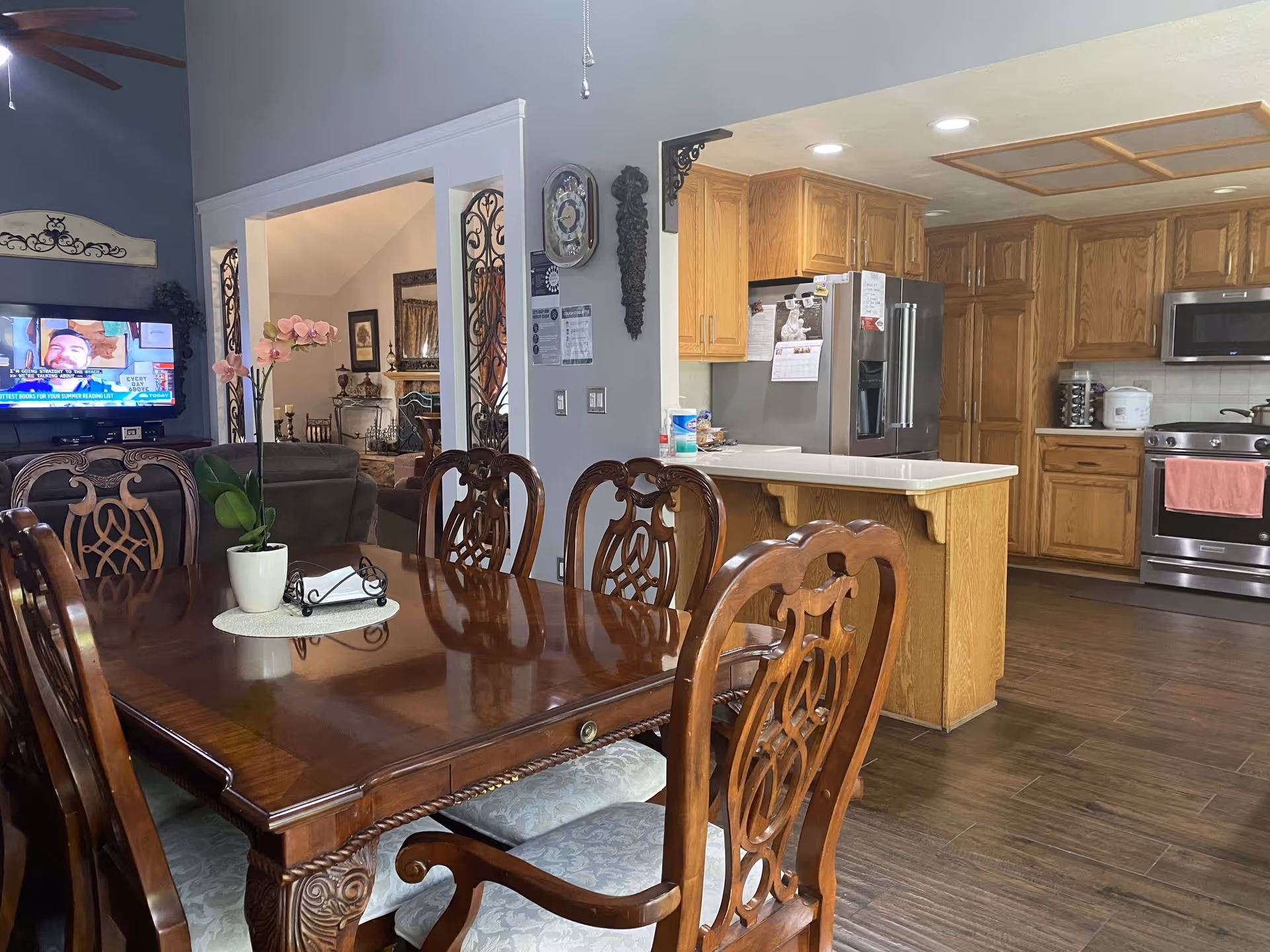 Ornate wooden dining table with a potted orchid and carved chairs in an open-plan dining area opening to a kitchen with oak cabinets and stainless steel appliances.