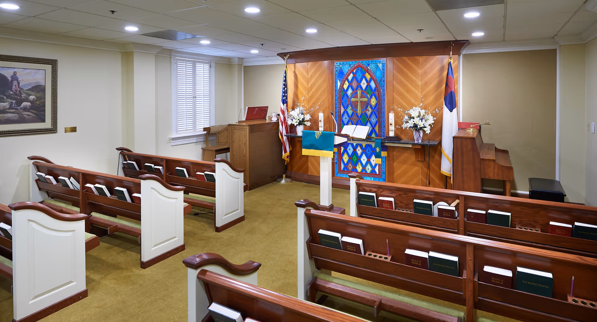 Interior view of a small chapel or worship room with wooden pews, hymnals, and Bibles placed in holders. At the front, there is a lectern with a green cloth, two flower arrangements, an American flag, a Christian flag, a piano, and a stained glass window featuring a cross design.