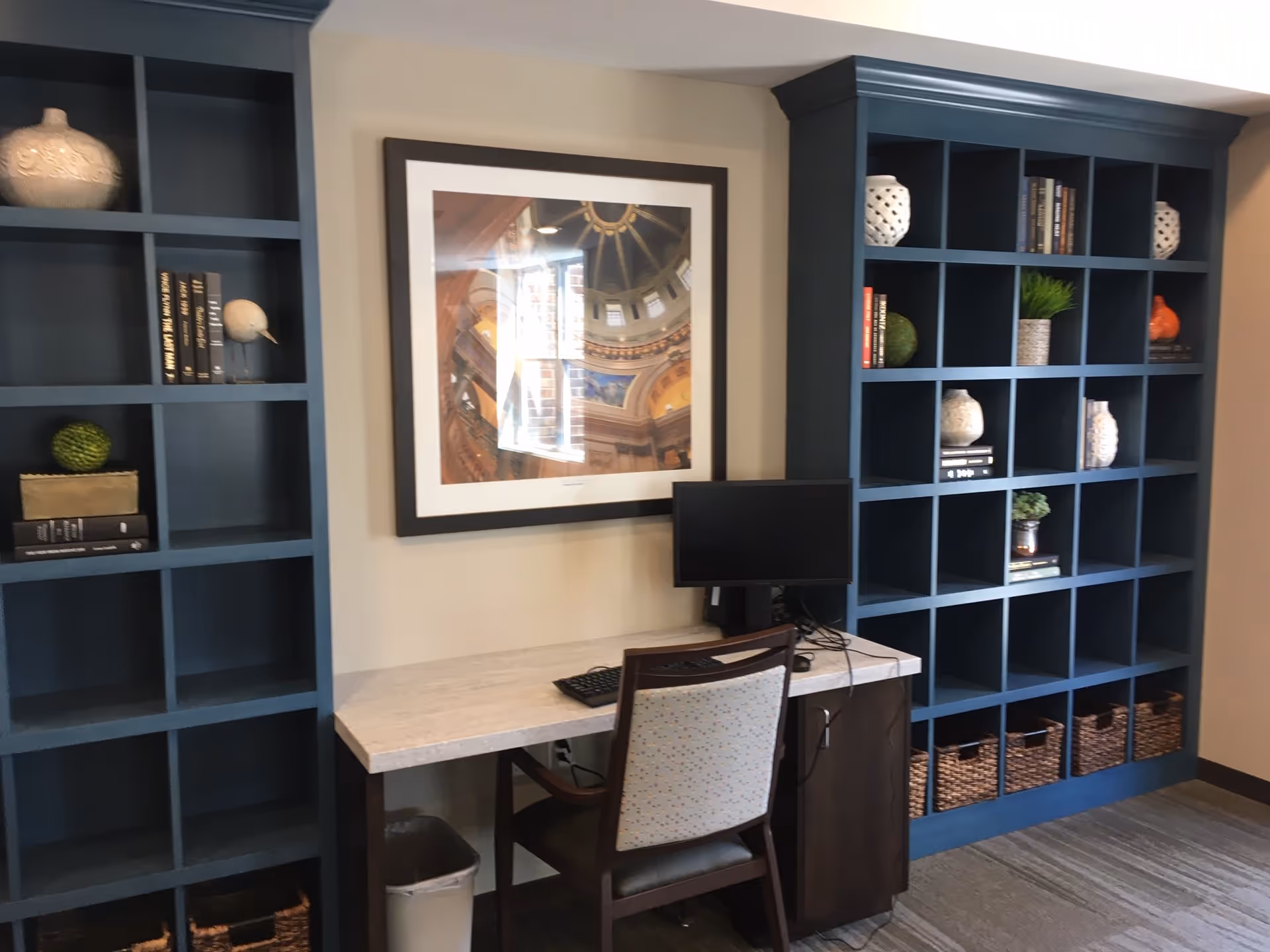 A small office or study area with a computer on a desk, a chair, and built-in blue bookshelves on either side. The shelves contain decorative items such as vases, books, plants, and baskets. A framed picture hangs on the wall above the desk.