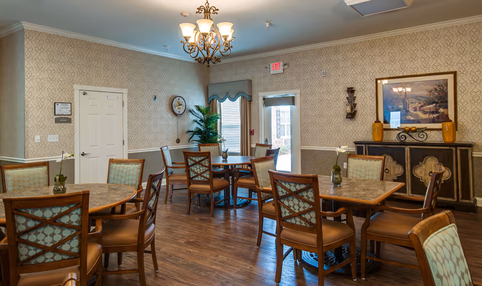 A well-lit dining room in a senior living facility with several round tables and wooden chairs with patterned cushions. The room features hardwood flooring, patterned wallpaper, a chandelier, a sideboard with decorative items, a framed painting, and a door leading outside with an exit sign above it.