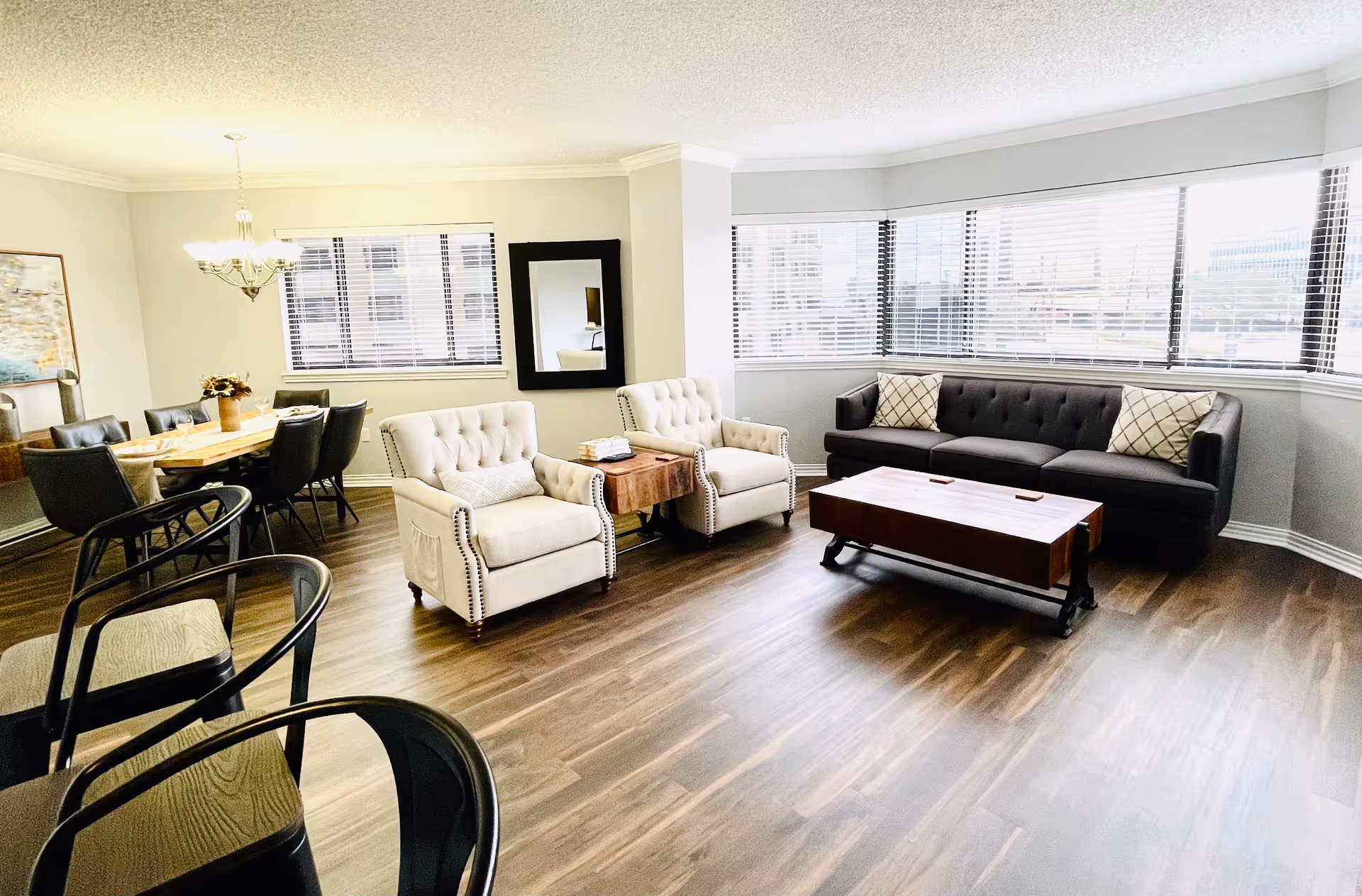 A bright and spacious living room area with large windows and wooden flooring. The room features a dark gray sofa with two patterned pillows, two beige armchairs with nailhead trim, a wooden coffee table, and a small side table with books. To the left, there is a dining area with a wooden table, six black chairs, a chandelier overhead, and a framed artwork on the wall.