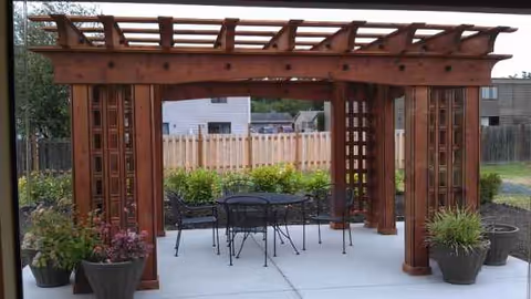 Outdoor wooden pergola with table, chairs, and potted plants on a patio.