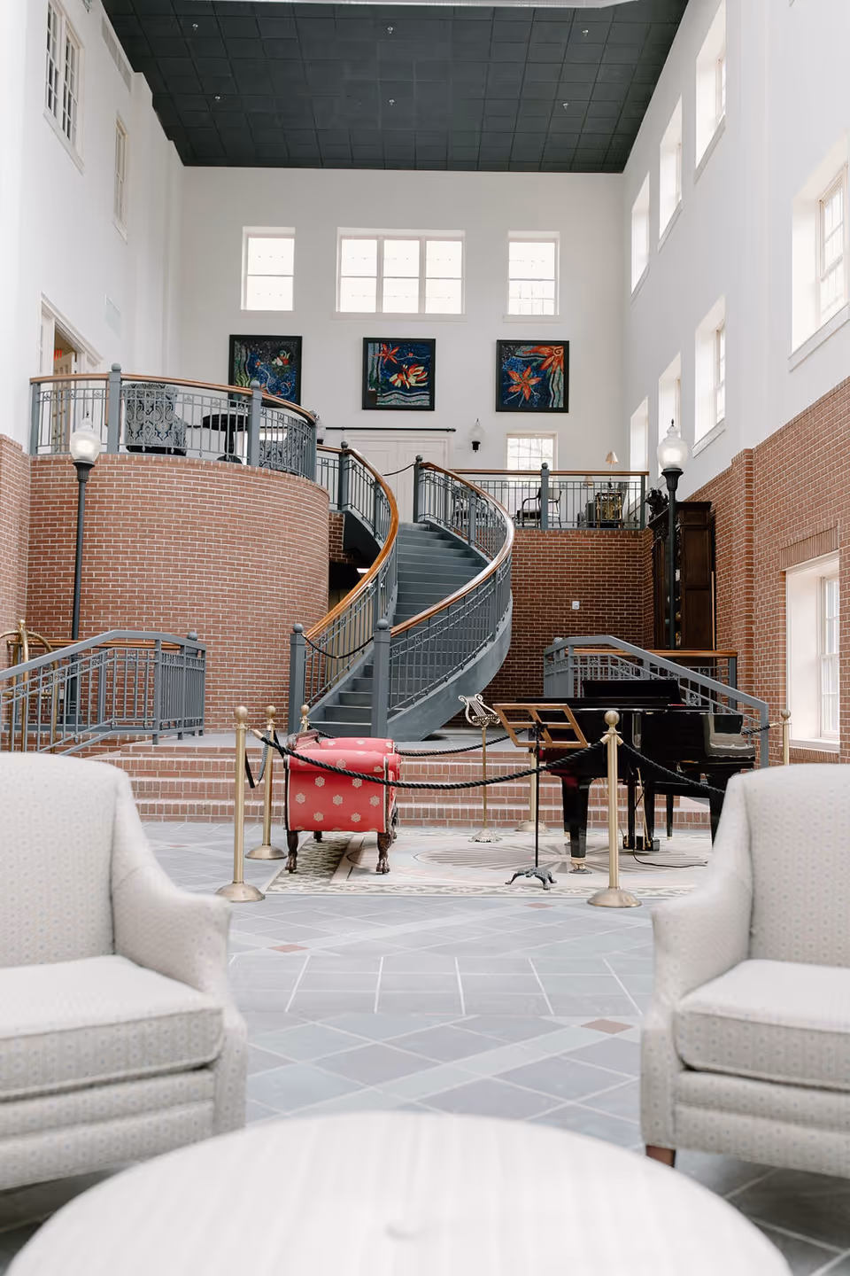 Spacious interior of The Madison Assisted Living featuring a curved staircase with metal railings and wooden handrails, brick walls, large windows, and three colorful paintings on the far wall. In the foreground, two light-colored upholstered armchairs face a round table, and a black grand piano is roped off near the staircase.