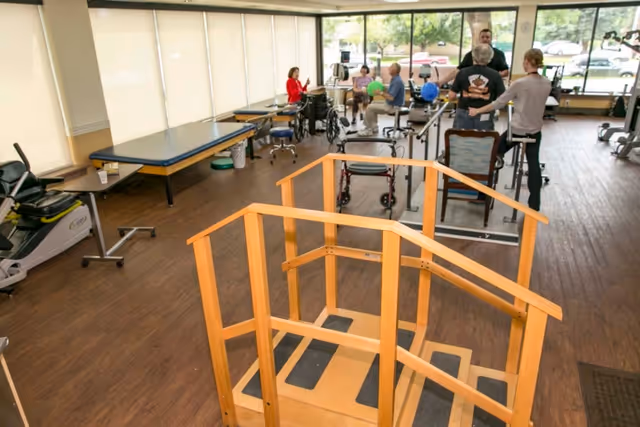 A rehabilitation or physical therapy room with exercise equipment, parallel bars, a wooden step training platform, and several people engaged in therapy activities. The room has large windows letting in natural light and wooden flooring.