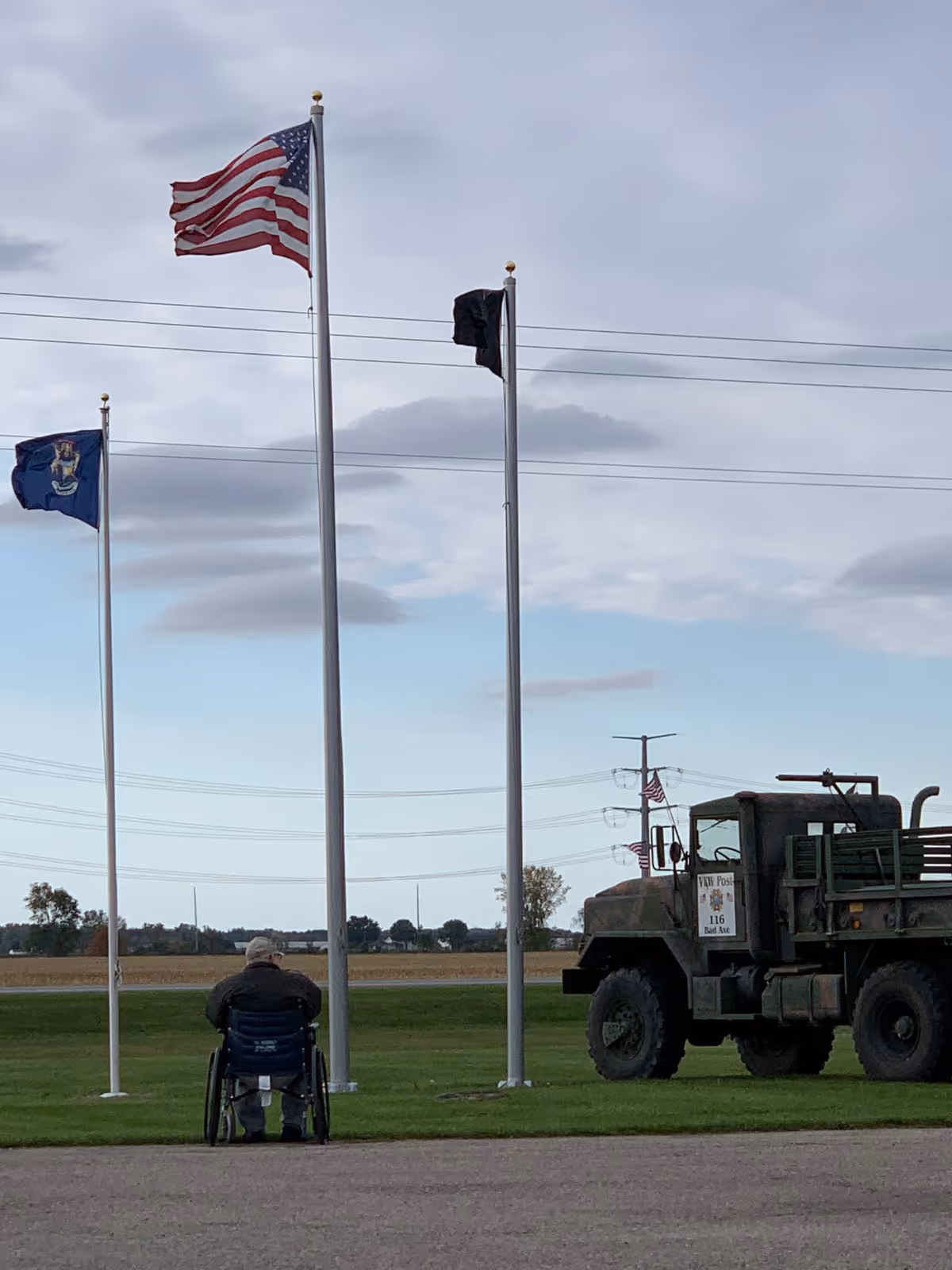 An elderly person in a wheelchair faces three flagpoles with flags and a military truck on a grassy field under a cloudy sky.