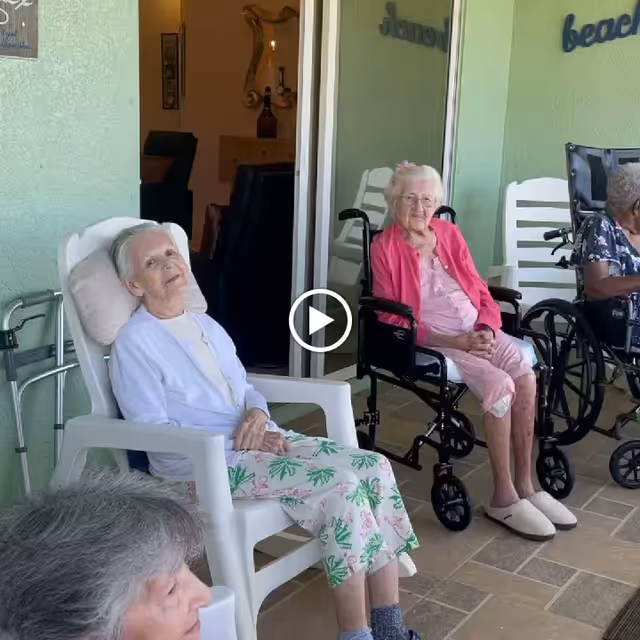 Several elderly women sitting outside on a patio area. One woman is seated in a white plastic chair wearing a light blue cardigan and floral pants, another woman in a wheelchair wearing a pink outfit and slippers, and another woman partially visible also in a wheelchair. The background shows a green wall with a sliding glass door and some decorative text on the wall.