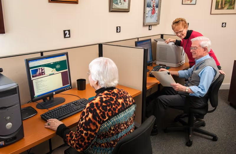 Two elderly individuals seated at separate computer stations in a room with cubicle dividers. One woman with white hair and a patterned sweater is using a desktop computer, while an elderly man in a blue shirt and gray vest is holding a clipboard and smiling. Another woman in a red sweater is standing behind the man, looking at his clipboard and smiling. The room has framed artwork on the walls and numbered cubicles.