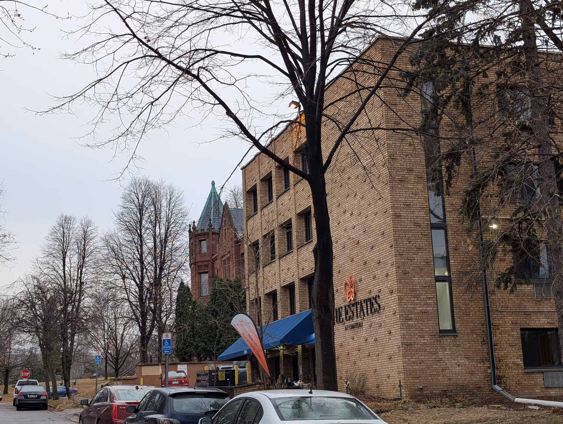 Brick multi-story senior living building displaying 'The Estates' signage with a blue awning, parked cars, and leafless trees in front.