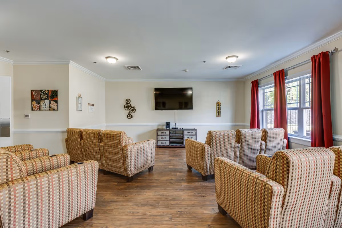A senior living lounge with patterned armchairs arranged facing a wall-mounted TV and large windows with red curtains.