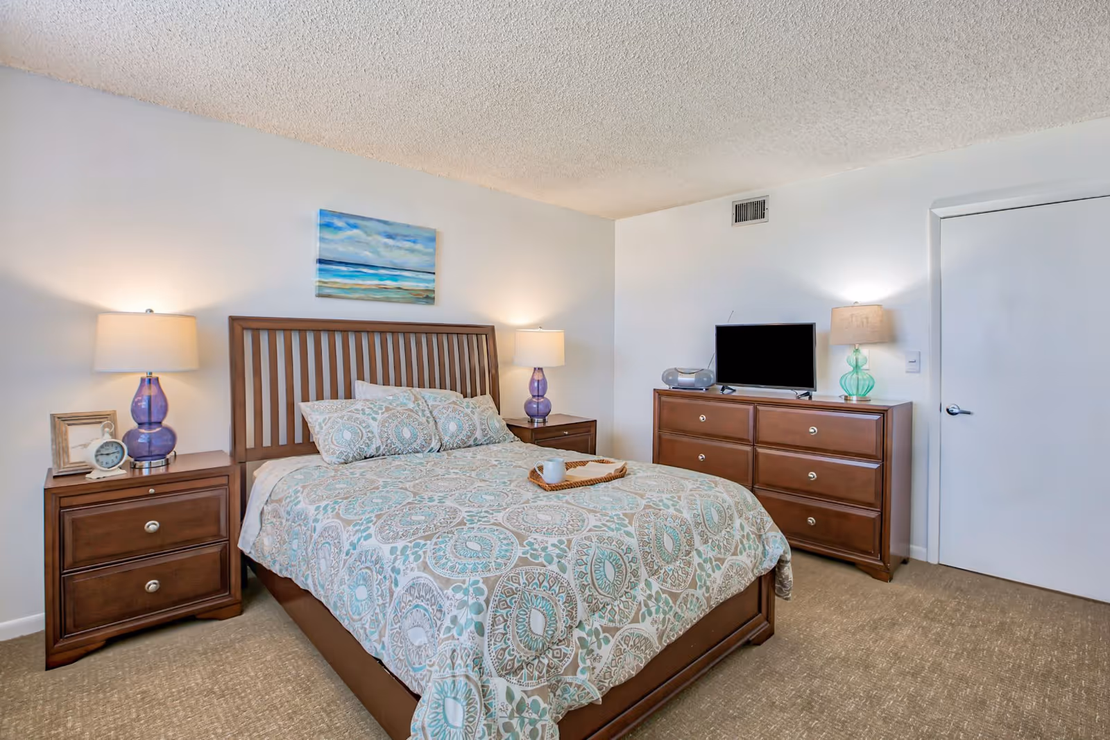 A bedroom with a wooden bed frame and a patterned bedspread in shades of blue and beige. There are two matching wooden nightstands with lamps on either side of the bed. A wooden dresser with a TV and a lamp is against the wall, and a door is visible on the right side of the room. A painting of a seascape hangs above the bed.