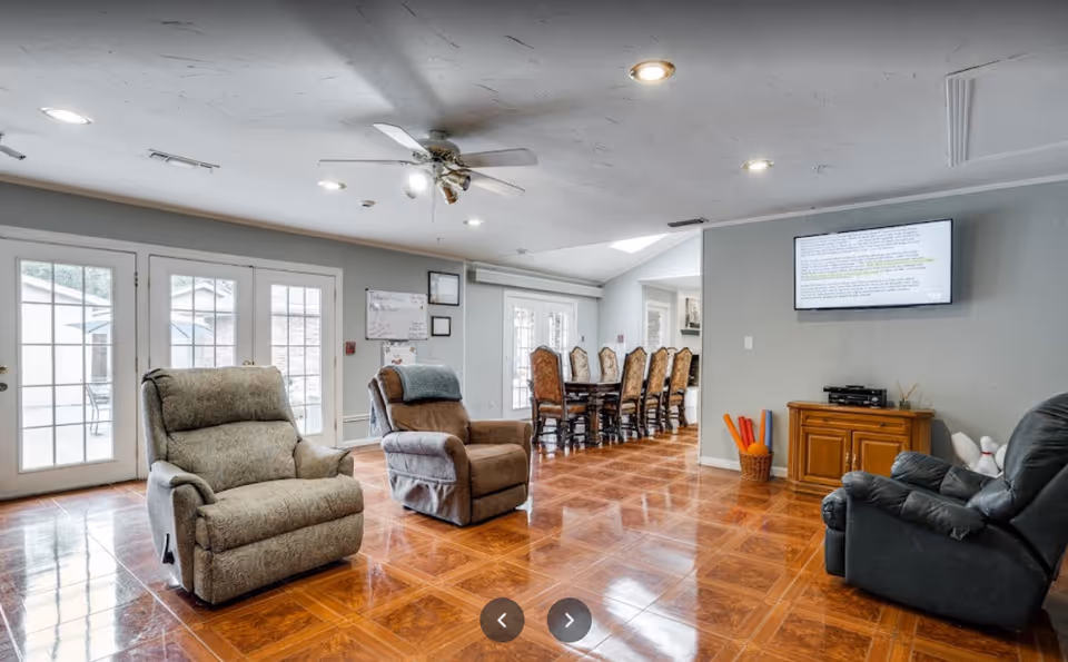 A spacious living area with three recliner chairs arranged on a polished wooden floor. In the background, there is a long dining table with multiple chairs. The room has large glass doors letting in natural light, a ceiling fan with lights, and a wall-mounted TV above a wooden cabinet. Various items like yoga mats and a bowling pin set are visible near the cabinet.