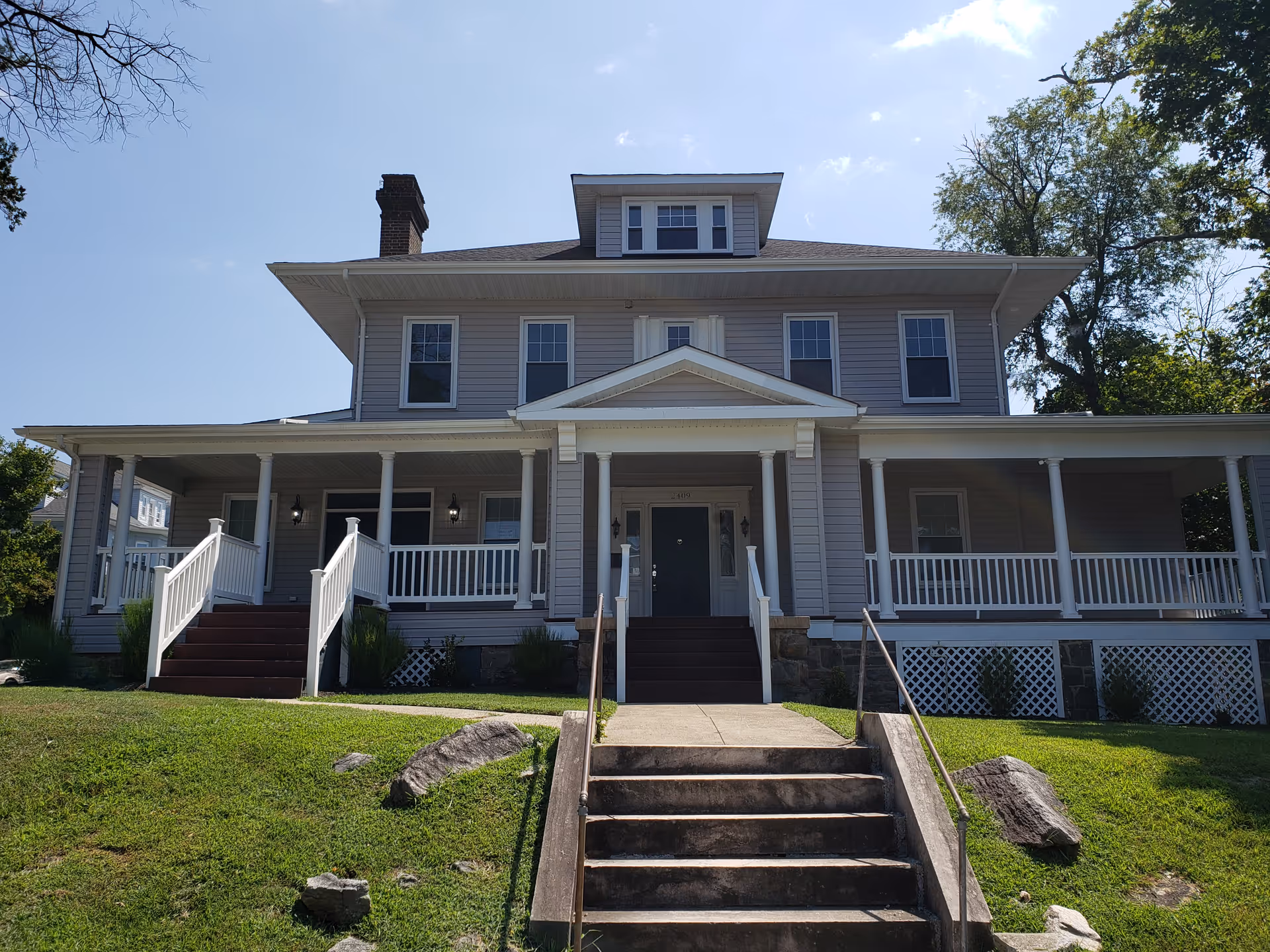 Front exterior view of a large two-story house with a wide porch, white railings, and steps leading up to the main entrance. The house is surrounded by a grassy lawn with some rocks and trees in the background under a clear blue sky.