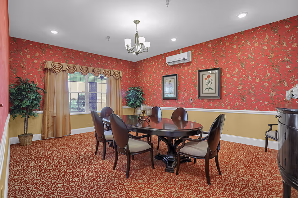A formal dining room with red and gold floral wallpaper, a patterned red carpet, and a large wooden dining table surrounded by six chairs. The room has a window with gold curtains, two framed botanical prints on the wall, two potted plants, a chandelier, and a wooden sideboard with a flower arrangement.