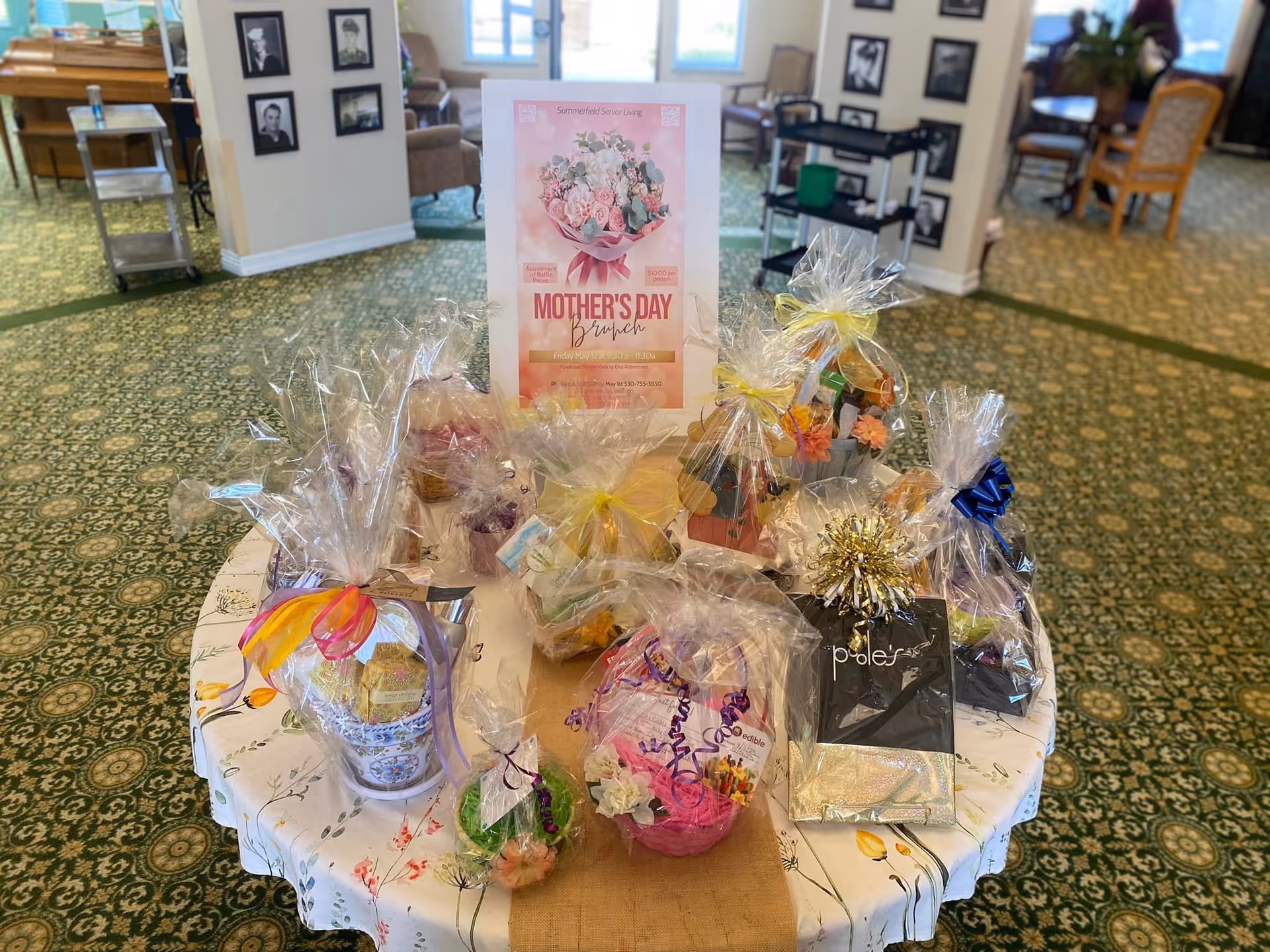 A round table in a senior living common area filled with wrapped Mother's Day gift baskets and a Mother's Day Brunch sign.