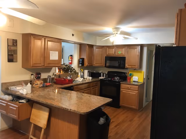 Interior view of a kitchen with wooden cabinets, a granite countertop island with a wooden chair, a black stove and microwave, a black refrigerator, and various kitchen items on the counters including a red dish rack and a fire extinguisher mounted on the wall.