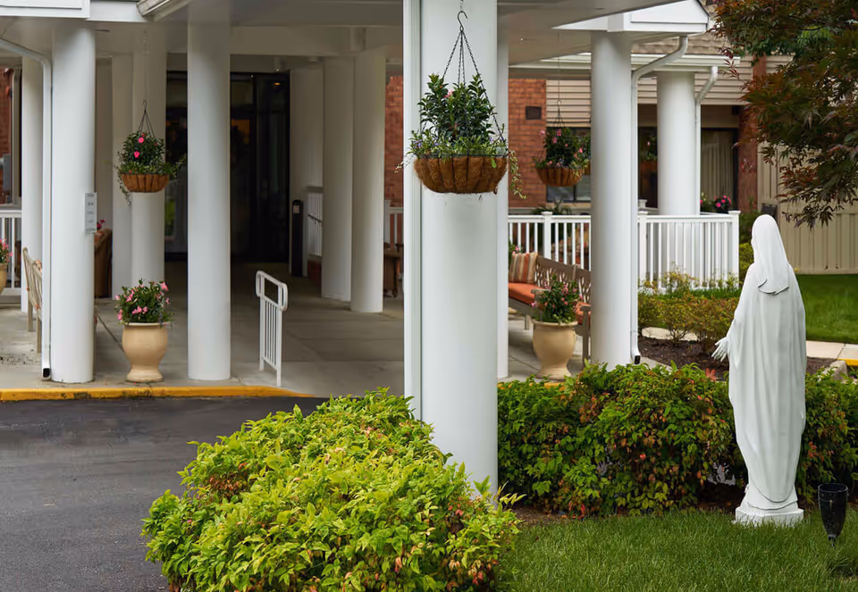 Covered building entrance with white columns, hanging flower baskets, potted plants, a white statue, and landscaped bushes.