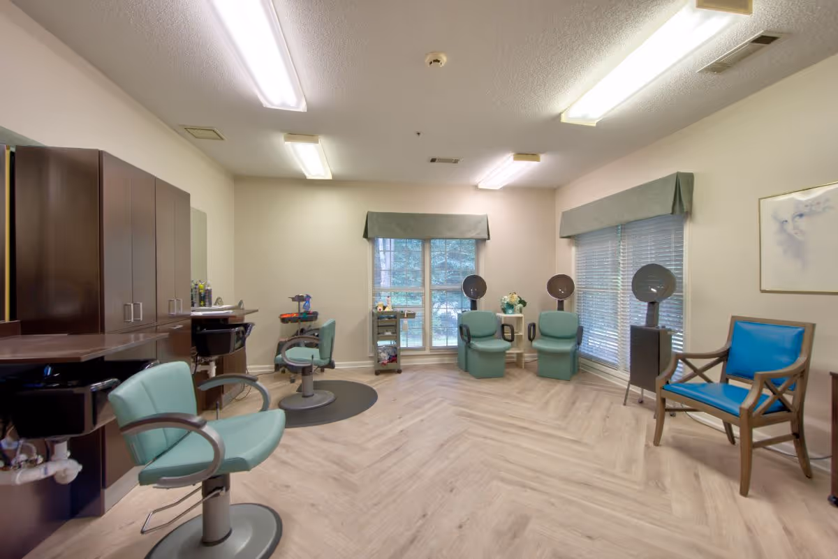 Interior of a salon room with light wood flooring, two green salon chairs with sinks, two green chairs with hair dryers, a blue cushioned wooden chair, large windows with blinds and valances, and overhead fluorescent lighting.