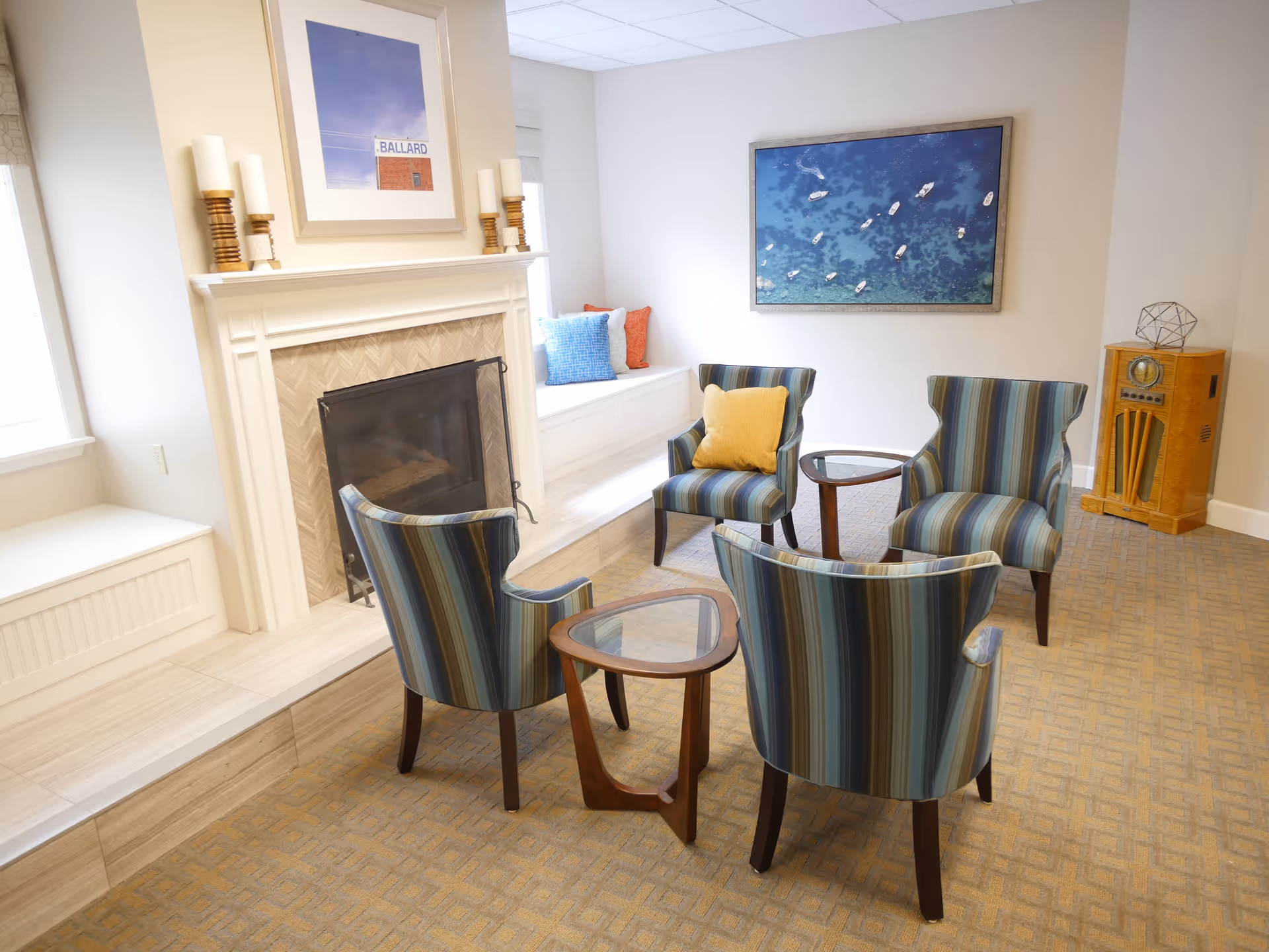 A bright lounge with four striped armchairs arranged around small tables facing a fireplace and coastal wall art.