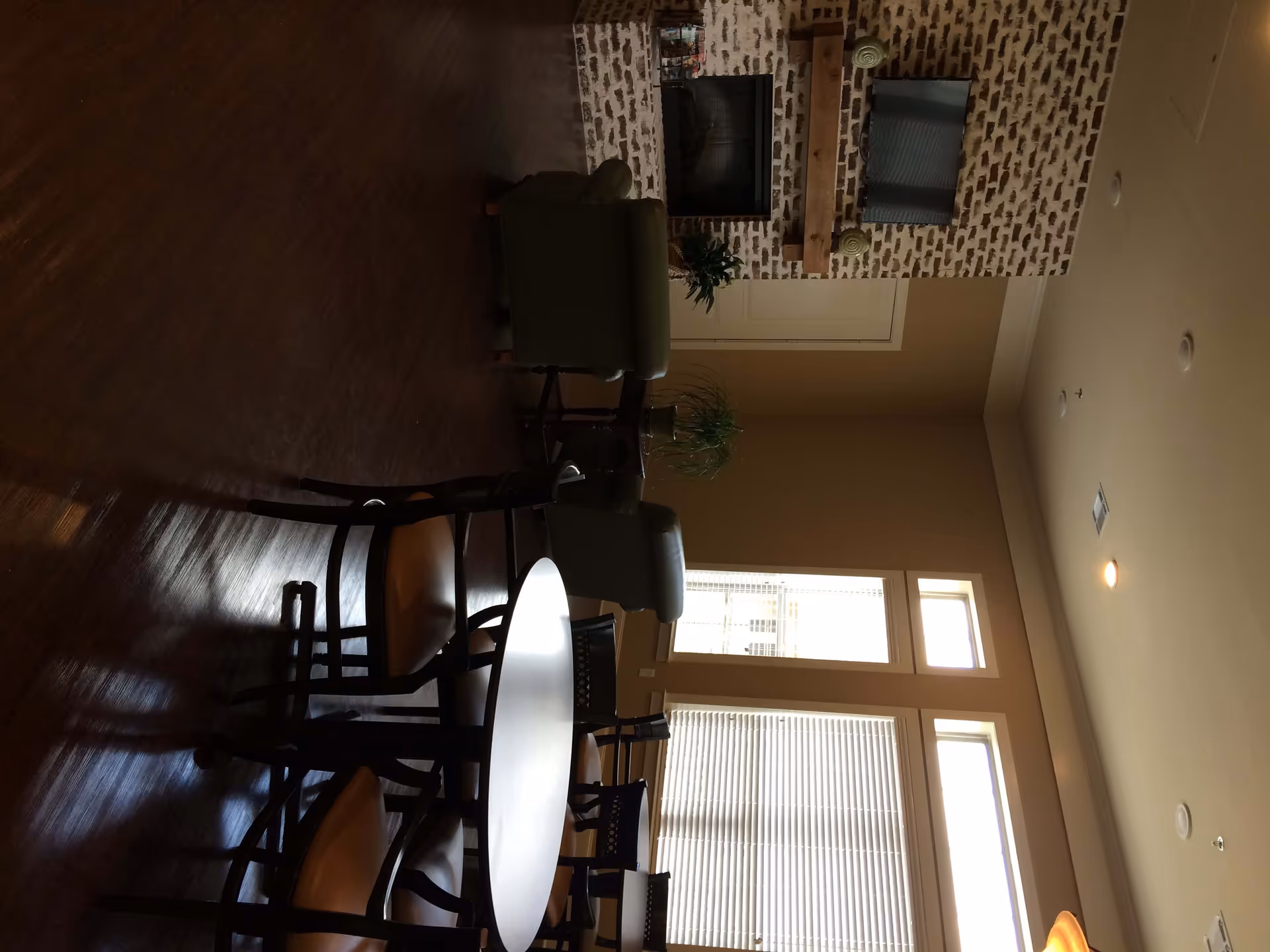 A cozy common area with a round table surrounded by chairs in the foreground. In the background, there are two armchairs facing a brick fireplace with a mounted TV above it. Large windows with blinds allow natural light to fill the room, and there are potted plants on the table and near the fireplace. The room has wooden flooring and beige walls.