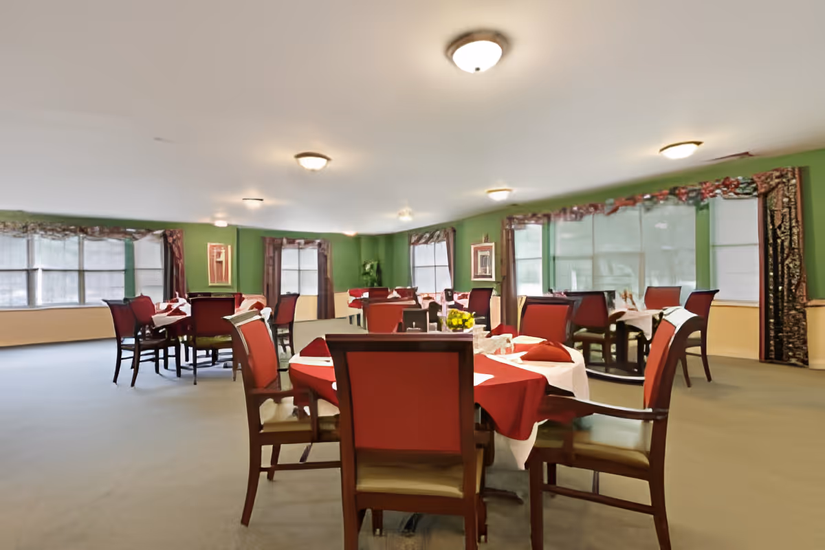 A spacious dining room with multiple tables covered in white and red tablecloths, surrounded by wooden chairs with red cushions. The walls are painted green with large windows covered by curtains, and ceiling lights illuminate the room.