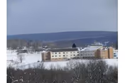 A large nursing center building situated in a snowy landscape with leafless trees in the foreground and hills in the background under a cloudy sky.
