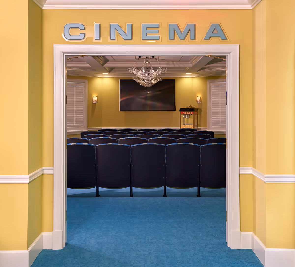 View through a doorway into a small cinema room with rows of black chairs facing a large flat screen TV mounted on a yellow wall. A popcorn machine is visible to the right of the screen, and a chandelier hangs from the ceiling. The word 'CINEMA' is displayed above the doorway.