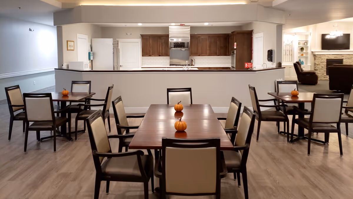 A dining area in an assisted living facility with several wooden tables and cushioned chairs arranged neatly. Small decorative pumpkins are placed on each table. In the background, there is a kitchen area with wooden cabinets, a refrigerator, and a stove. To the right, a living room area with armchairs and a TV mounted above a stone fireplace is visible.