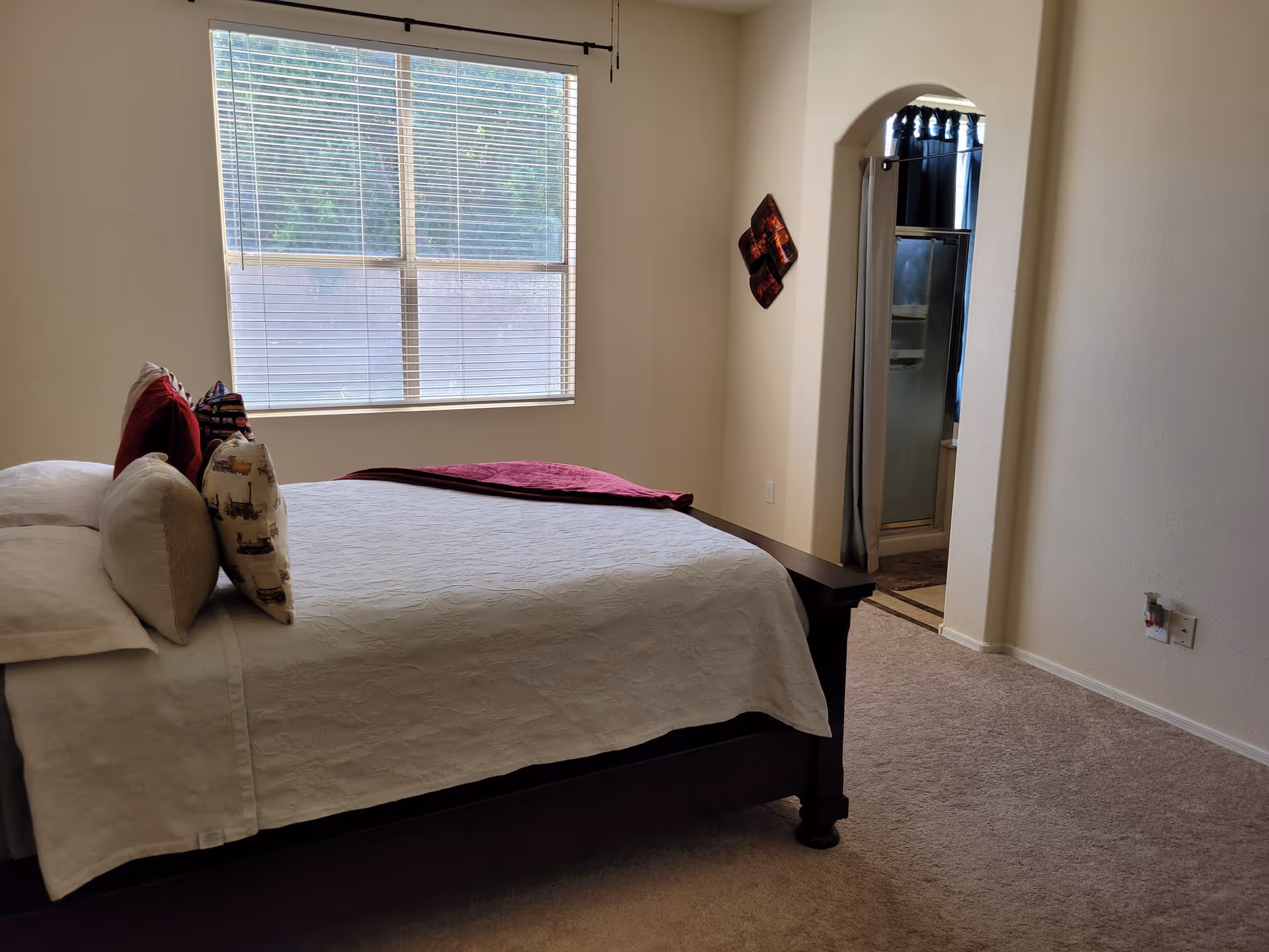 A bedroom with a large window covered by blinds, a bed with white bedding and multiple pillows, and an open doorway leading to a bathroom with a shower stall visible.