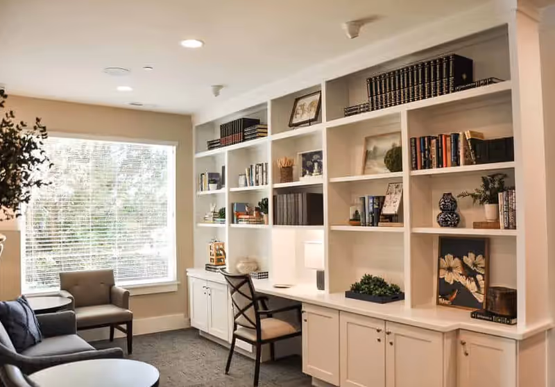 A cozy living room area with a large window letting in natural light, a gray sofa, a gray armchair, a round coffee table, and a built-in white bookshelf filled with books, framed pictures, and decorative items. A desk with a chair and a table lamp is integrated into the bookshelf.
