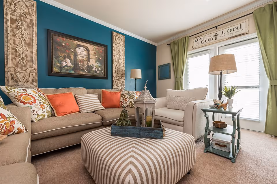 A cozy living room with a beige sectional sofa adorned with floral and orange cushions, a white armchair, and a striped ottoman in the center. The walls feature teal and beige colors with decorative wall art. A small green side table holds a lamp and decorative items next to large windows with green curtains and a sign above that reads 'Love the Lord your God with all your heart.'