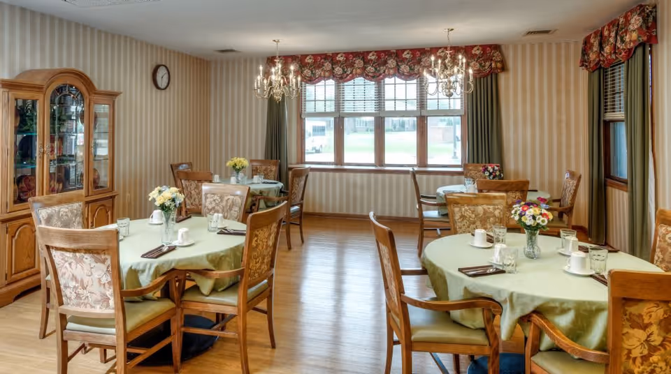 A dining room with several round tables covered with light green tablecloths, each set with cups, glasses, napkins, and floral centerpieces. The room has wooden chairs with floral upholstery, a wooden china cabinet, striped wallpaper, two chandeliers, and large windows with floral valances and green curtains.