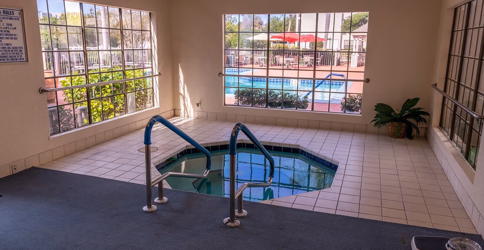 Indoor hot tub with metal handrails surrounded by tiled floor and large windows showing an outdoor pool area with tables, chairs, and red umbrellas.