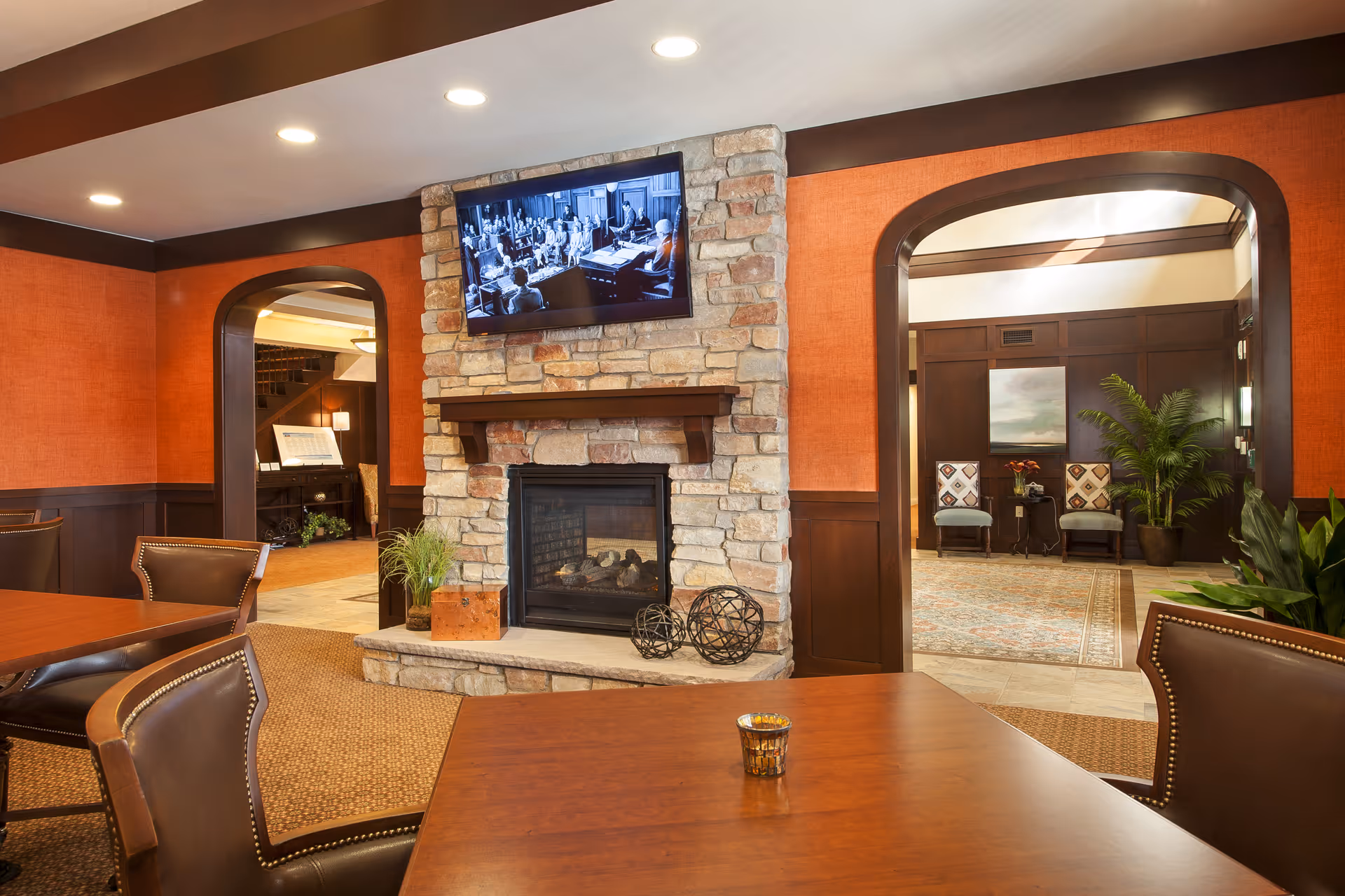 A cozy interior common area with a stone fireplace and a mounted flat-screen TV above it. The room features warm orange walls with dark wood trim, leather chairs around wooden tables, and two arched doorways leading to other rooms with additional seating and plants.