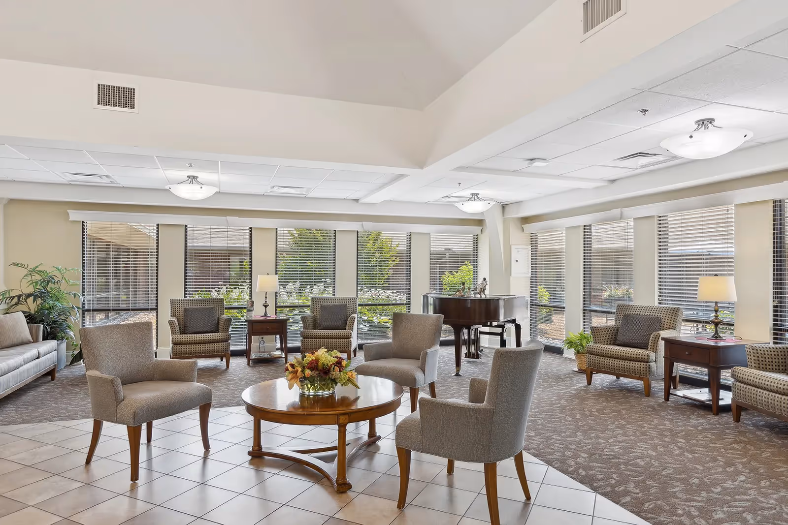 A bright and spacious living room area with multiple armchairs and a round wooden coffee table with a flower arrangement in the center. Large windows with blinds allow natural light to fill the room. There is a grand piano in the background and several side tables with lamps. The room has a mix of tiled and carpeted flooring and some indoor plants near the windows.