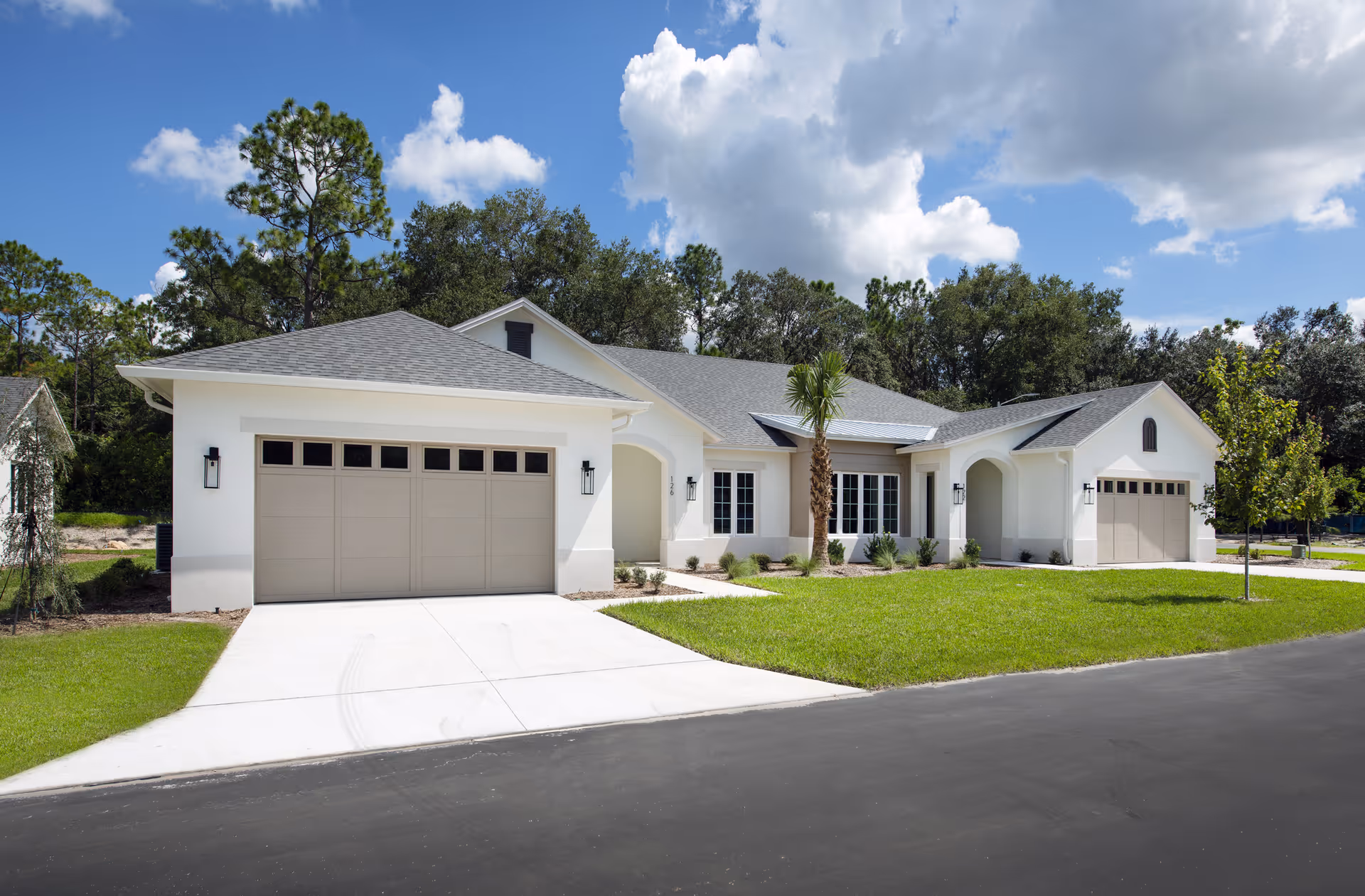 A single-story residential building with two beige garage doors, white exterior walls, and a gray shingled roof. The house is surrounded by a well-maintained green lawn, a few small trees, and shrubs. The sky is blue with scattered clouds.