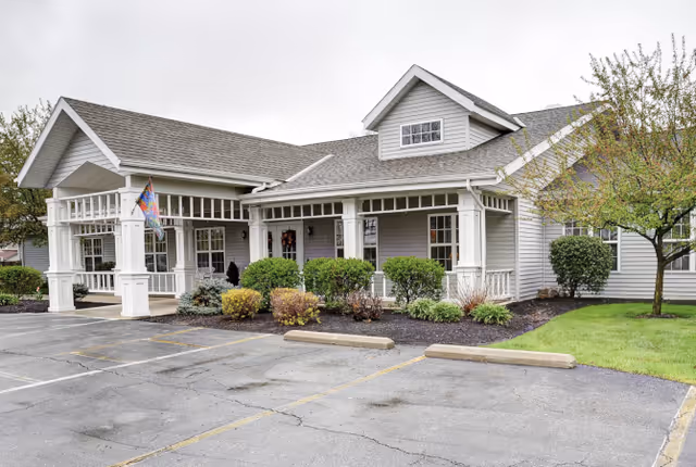 Front exterior of a single-story senior living facility with a covered entryway, white columns, landscaping, and an empty parking area.