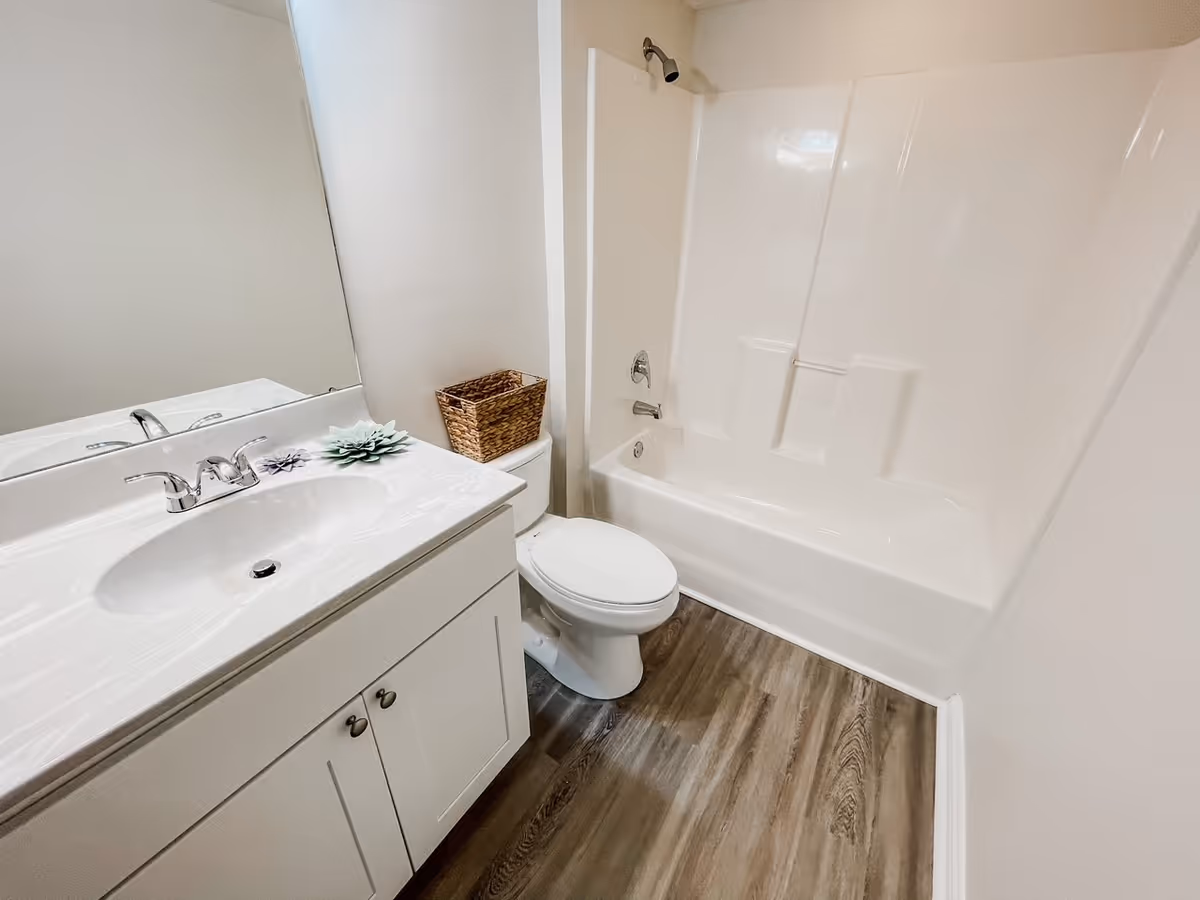 A clean bathroom featuring a white vanity with a sink and chrome faucet, a large mirror above the sink, a white toilet with a wicker basket on top, and a white bathtub with a showerhead. The floor has wood-like vinyl flooring and the walls are painted white.