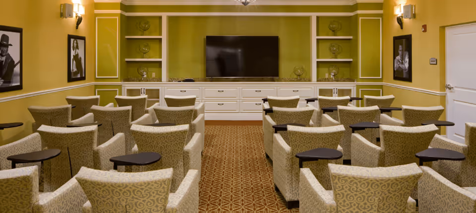 A small theater-style room with multiple rows of beige patterned armchairs, each equipped with a small black side table. The room has yellow walls with white trim, black and white framed photos on the walls, and a large flat-screen TV mounted on a green accent wall with built-in white cabinetry below and shelves on either side.